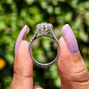 May include: A close-up of a silver ring with a large, clear, round-cut gemstone. The ring has a halo of smaller stones around the main stone and along the band. The ring is held between two fingers with a blurred green background.