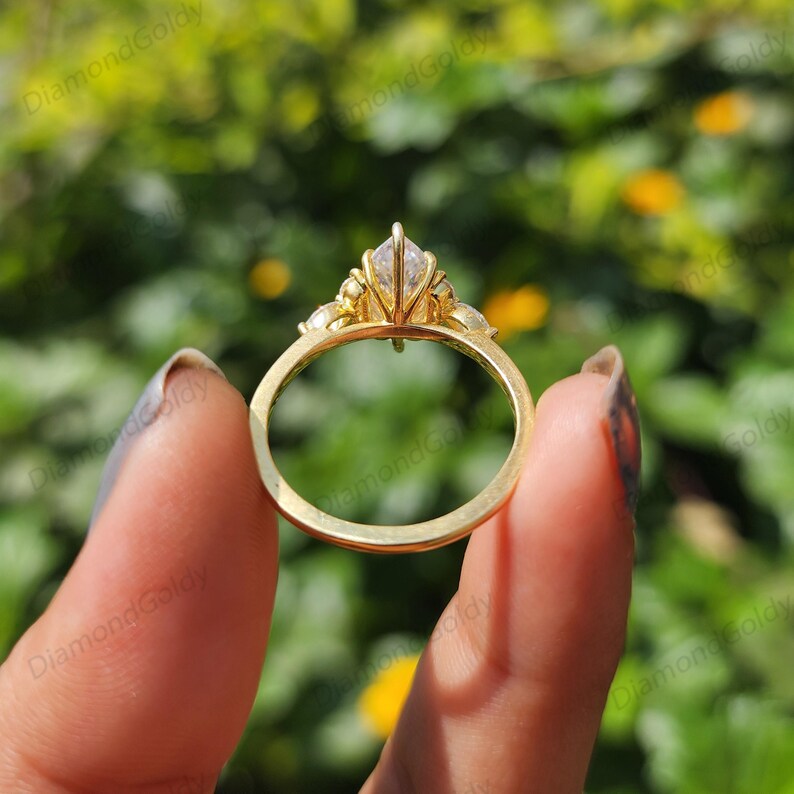 May include: A gold ring with a marquise-cut diamond center stone and smaller accent stones. The ring is held up against a blurred green background, showcasing its intricate design and sparkle. The ring's band is a simple, polished gold.