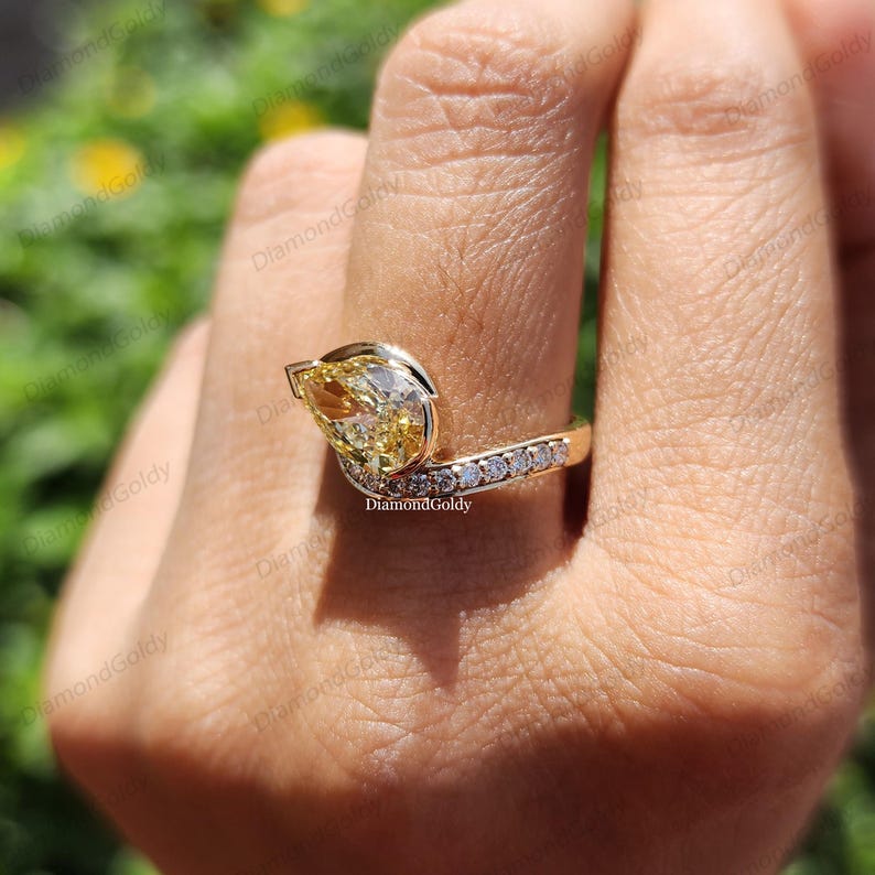 May include: A gold ring featuring a pear-shaped yellow diamond as the primary stone. The ring has a curved design with a row of smaller, clear diamonds set along the band. The ring is on a finger.