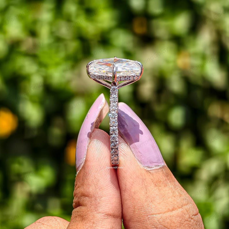 May include: A close-up of a diamond ring with a large, oval-shaped center stone. The ring has a halo of smaller diamonds around the center stone and a band lined with diamonds. The ring is set in a silver metal.