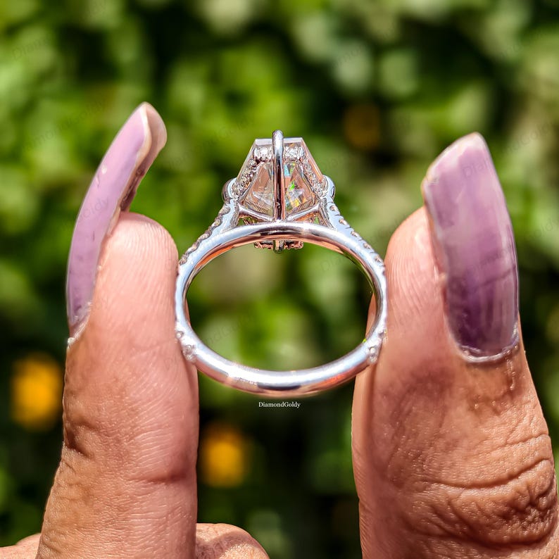May include: Close-up of a silver ring with a large, clear, emerald-cut center stone. The ring features a halo of smaller stones around the center and along the band. The ring is held up against a blurred green background.