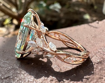 May include: Rose gold ring featuring a large, light green gemstone with a pear-cut shape. The ring is adorned with smaller, clear gemstones and a delicate, leaf-like design. The words "Diamond Goldy" are visible on the ring.