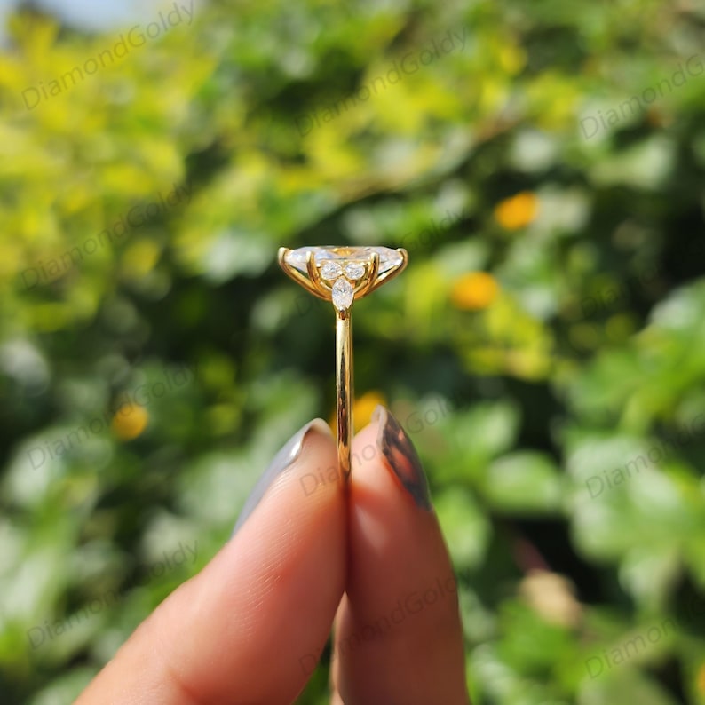 May include: A gold ring with a large, pear-shaped, clear gemstone as the centerpiece. The ring has smaller, clear gemstones on either side of the main stone. The ring is held up against a blurred green background.