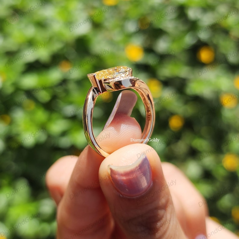 May include: A gold ring with a large, yellow, pear-shaped gemstone. The ring has a split shank design and is held up by a person's fingers. The background is blurred with green foliage and yellow flowers.