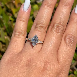 May include: A gold ring featuring a marquise-cut, light blue gemstone as the centerpiece, flanked by smaller, round, clear gemstones. The ring is set on a hand with white nail polish.