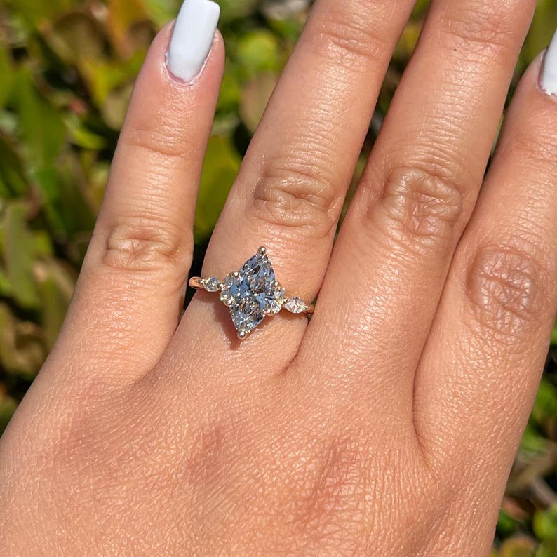 May include: A gold ring featuring a marquise-cut, light blue gemstone as the centerpiece, flanked by smaller, round, clear gemstones. The ring is set on a hand with white nail polish.