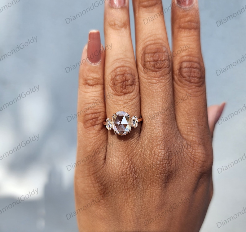 Op de afbeelding: Een ros&eacute;gouden ring met een grote, ovaal geslepen diamant als middensteen, geflankeerd door twee kleinere, ronde diamanten. De ring wordt getoond op een hand met een lichte huidskleur, waardoor de schittering van de edelstenen wordt benadrukt. Een sieraad.