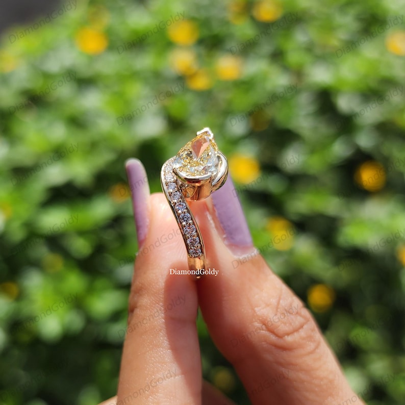 May include: A gold ring with a pear-shaped yellow gemstone and a curved band. The band is adorned with small, clear gemstones. The ring is held up against a blurred green and yellow background. The text "DiamondGoldy" is visible.