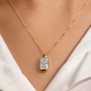 May include: A gold chain necklace featuring a rectangular radiant-cut clear gemstone pendant set in a gold frame. The pendant hangs vertically from a delicate gold link chain, resting against a white fabric surface with a visible watermark pattern.