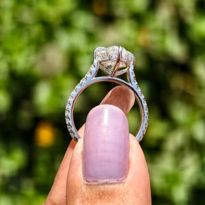 May include: A silver engagement ring with a large, clear, cushion-cut diamond center stone. The band is encrusted with smaller diamonds. The ring is held up against a blurred green background.