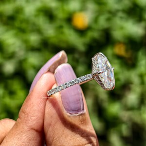 May include: A silver engagement ring with a large, clear, oval-cut diamond as the centerpiece. The band is encrusted with small, sparkling diamonds. The ring is held up against a blurred green background.