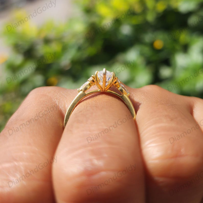 May include: A gold engagement ring with a central, clear, pear-shaped gemstone, flanked by smaller, rectangular stones. The ring's band is a simple, polished gold. The ring is held by a hand, with a blurred green background.