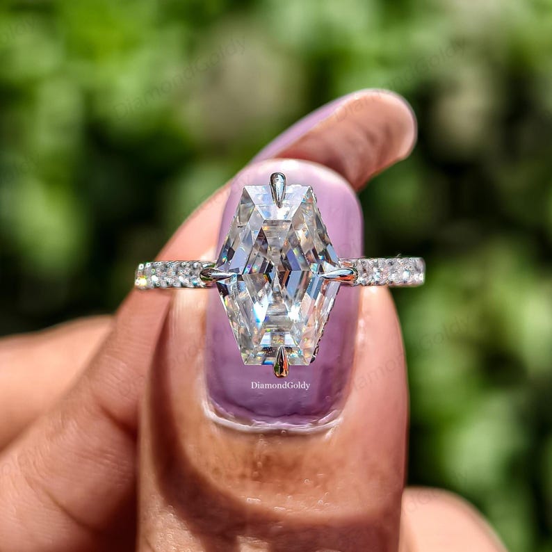 May include: A close-up of a diamond ring with a unique, geometric cut. The large, clear diamond is the focal point, set on a band with smaller, sparkling stones. The ring is held against a blurred green background.
