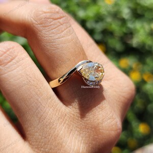 May include: A gold ring with a large, oval-shaped yellow gemstone. The ring features a unique design with a curved band and small accent diamonds. The ring is shown on a finger, with a blurred background.