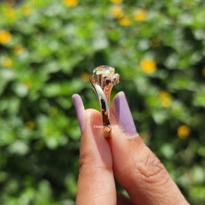 May include: A gold-colored ring with a large, light-colored gemstone and smaller accent stones. The ring has a unique, swirling design and is held up against a blurred green and yellow background.
