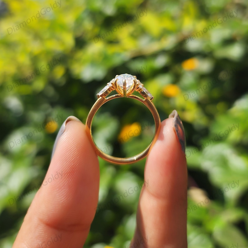 May include: A gold engagement ring with a large, central, pear-shaped diamond and two smaller side stones. The ring is held up against a blurred green background, showcasing its intricate design and sparkle.