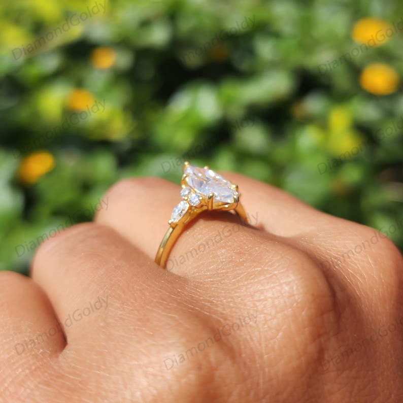 May include: A gold ring featuring a pear-shaped diamond center stone and smaller accent stones. The ring's band and setting are gold, and the stones sparkle against a blurred green background. The ring is shown on a person's finger.
