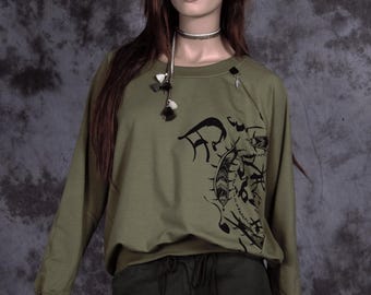 May include: A green long-sleeved sweatshirt with a black graphic print on the front. The sweatshirt has a zipper detail on the sleeves and a drawstring waistband. The model is wearing the sweatshirt with dark green cargo pants.
