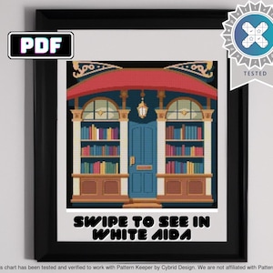 Op de afbeelding: Ingelijst kruissteekpatroon van een gebouw met een rood luifel, blauwe deur en boekenkasten. De afbeelding bevat de tekst "PDF" en "SWIPE TO SEE IN WHITE AIDA". Een "TESTED"-badge staat in de rechterbovenhoek.