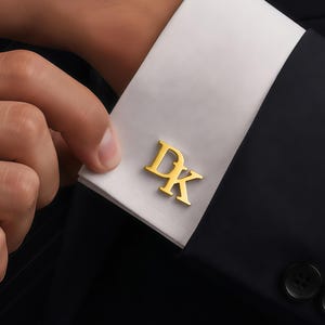 May include: A gold-toned metal cufflink featuring the letters D and K in a serif font, attached to a crisp white French cuff of a shirt sleeve. The cuff is worn under a dark navy blue blazer with visible black buttons on the sleeve cuff.