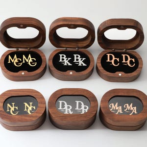 May include: Six oval-shaped dark wood boxes, each containing a pair of metallic cufflinks. The cufflinks feature various two-letter monograms in gold, silver, and rose gold tones. The boxes are arranged in two rows of three, with lids open or closed.