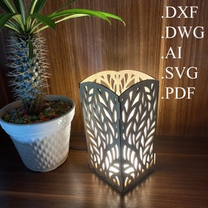 Op de afbeelding: Een houten lamp met een lichtbruine afwerking en een uitgesneden ontwerp van bladeren. De lamp is aan en geeft een warm licht. De lamp staat op een houten oppervlak. De tekst .DXF .DWG .AI .SVG .PDF is zichtbaar in de rechterbovenhoek van de afbeelding.
