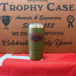 May include: A green, stainless steel tumbler with a brown football design and the word "Football" on it. The tumbler is sitting on a red, white, and blue surface. The background is a brown leather-like surface with the text "The Trophy Case" and "Awards & Engraving" on it.