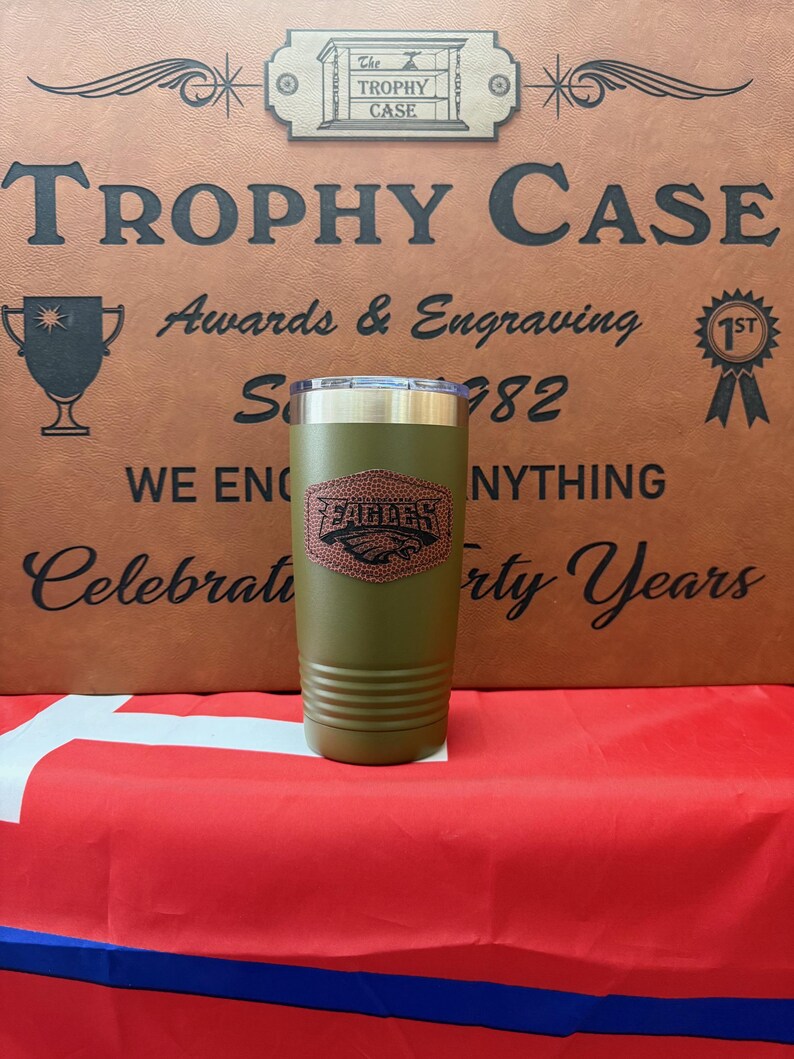 May include: A green, stainless steel tumbler with a brown football design and the word "Football" on it. The tumbler is sitting on a red, white, and blue surface. The background is a brown leather-like surface with the text "The Trophy Case" and "Awards & Engraving" on it.