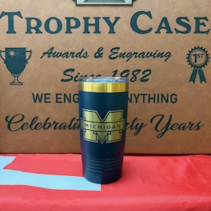 May include: A navy blue tumbler with a gold rim and the University of Michigan logo. The tumbler is set against a background that reads "The Trophy Case" and "Celebrating Sixty Years."