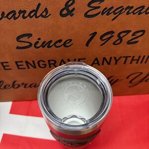May include: A red and stainless steel travel mug with a black leather-like band around the middle. The lid is silver and has a black logo that says "Polar Camel" with a mountain and a camel silhouette.