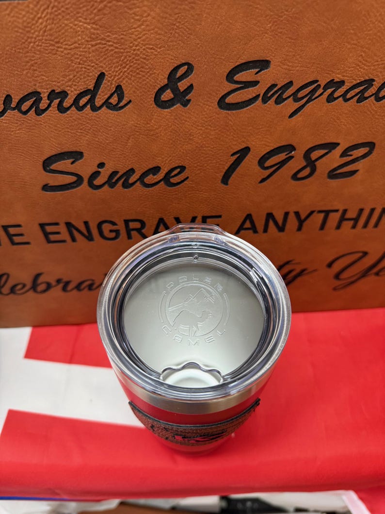 May include: A red and stainless steel travel mug with a black leather-like band around the middle. The lid is silver and has a black logo that says "Polar Camel" with a mountain and a camel silhouette.