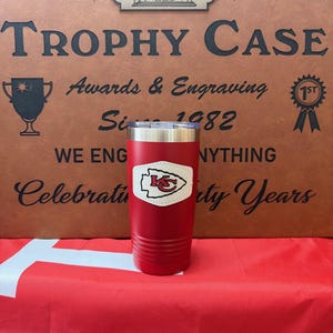 May include: Red stainless steel tumbler with a white leather patch featuring the Kansas City Chiefs logo. The tumbler is sitting on a red and blue surface in front of a brown sign that reads "The Trophy Case Awards & Engraving Since 1982".
