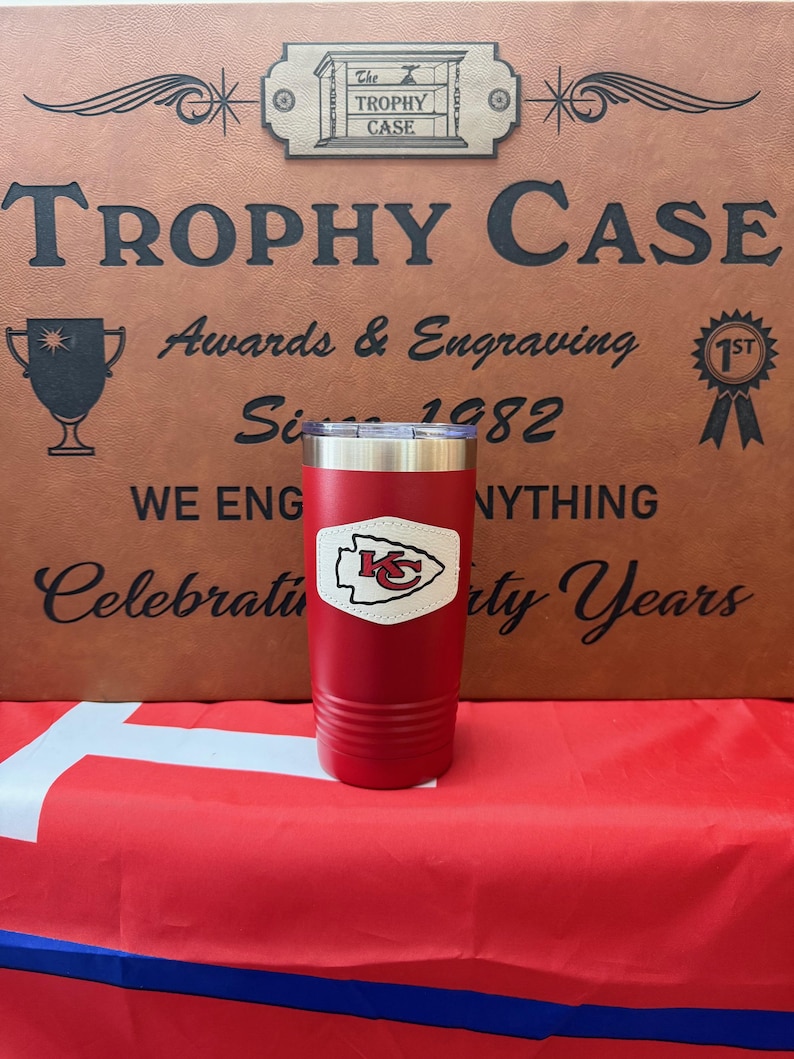 May include: Red stainless steel tumbler with a white leather patch featuring the Kansas City Chiefs logo. The tumbler is sitting on a red and blue surface in front of a brown sign that reads "The Trophy Case Awards & Engraving Since 1982".