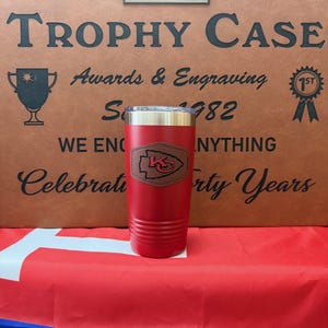 May include: Red stainless steel tumbler with a brown leather patch featuring a team logo. The tumbler is on a red, white, and blue surface. The text "The Trophy Case" is visible in the background.