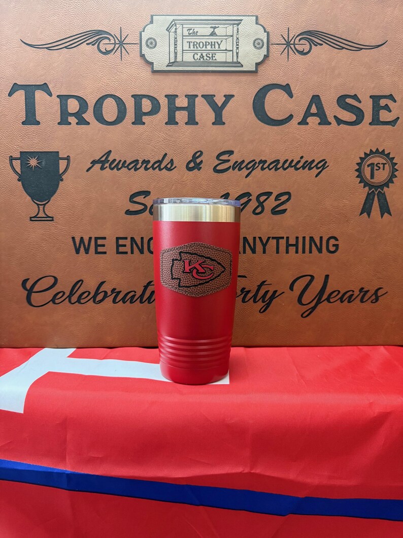 May include: Red stainless steel tumbler with a brown leather patch featuring a team logo. The tumbler is on a red, white, and blue surface. The text "The Trophy Case" is visible in the background.