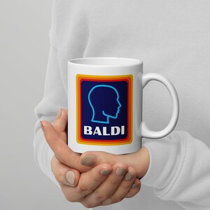 May include: White ceramic coffee mug with a colorful graphic of a blue head silhouette and the word "BALDI" in a rainbow font.