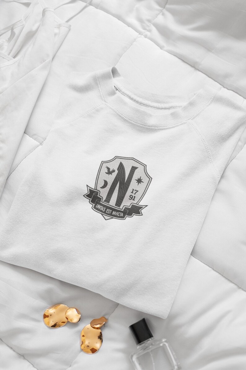 May include: White sweatshirt with a gray crest featuring a stylized letter N, a crescent moon, a star, a bird, and the text "unitas est invicta". The crest also includes the numbers 17 and 91.