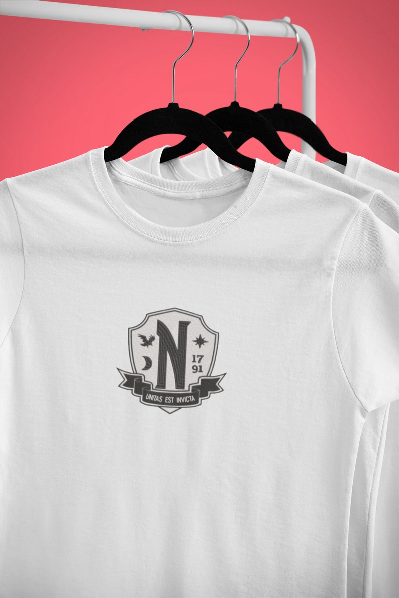 May include: White t-shirt with a black and gray crest featuring a stylized "N", a crescent moon, a star, a bird, and the text "17 91 UNITAS EST INVICTA".