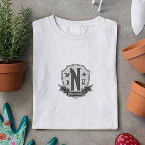 May include: White t-shirt with a gray crest featuring a stylized letter "N", a crescent moon, a star, and the text "unitas est invicta".