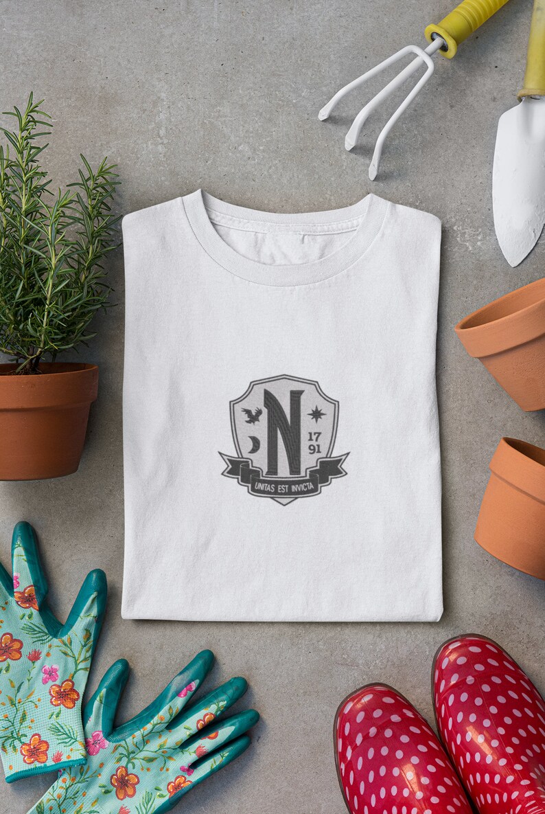 May include: White t-shirt with a gray crest featuring a stylized letter "N", a crescent moon, a star, and the text "unitas est invicta".