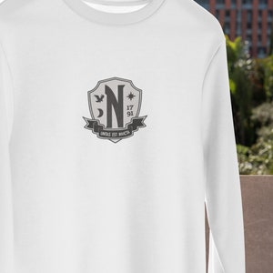 May include: White long-sleeve shirt with a gray crest featuring a stylized letter "N", a star, a crescent moon, and a bird. The crest also includes the numbers "17" and "91" and the text "UNITAS EST INVICTA".