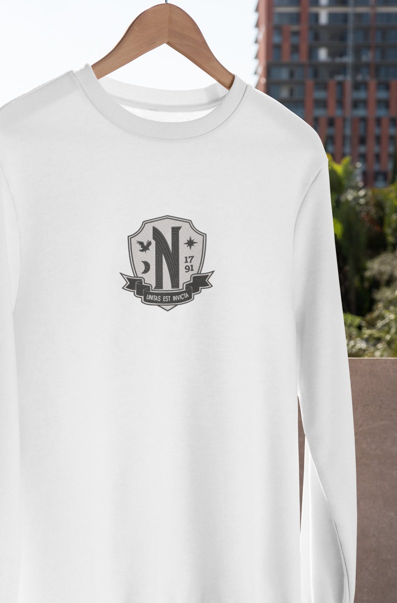 May include: White long-sleeve shirt with a gray crest featuring a stylized letter "N", a star, a crescent moon, and a bird. The crest also includes the numbers "17" and "91" and the text "UNITAS EST INVICTA".