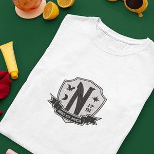 May include: White t-shirt with a black and white crest featuring a stylized letter N, a crescent moon, a star, and the text "Unitas Est Invicta". The crest also includes the numbers 17 and 91.