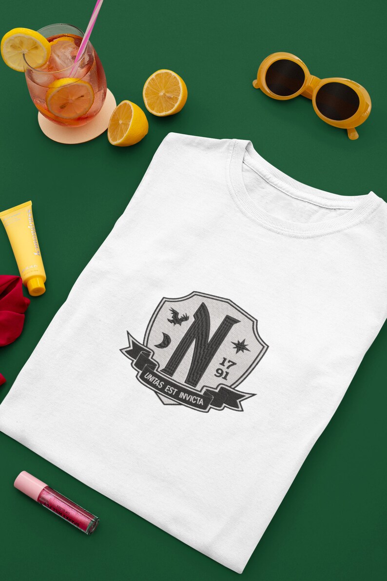 May include: White t-shirt with a black and white crest featuring a stylized letter N, a crescent moon, a star, and the text "Unitas Est Invicta". The crest also includes the numbers 17 and 91.