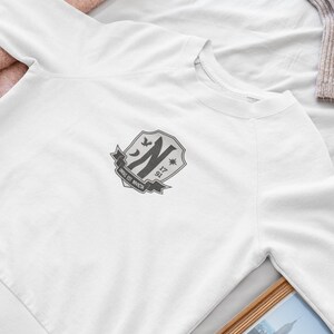 May include: White sweatshirt with a gray embroidered crest featuring a stylized letter "N", a star, and the text "NINE 17 91".