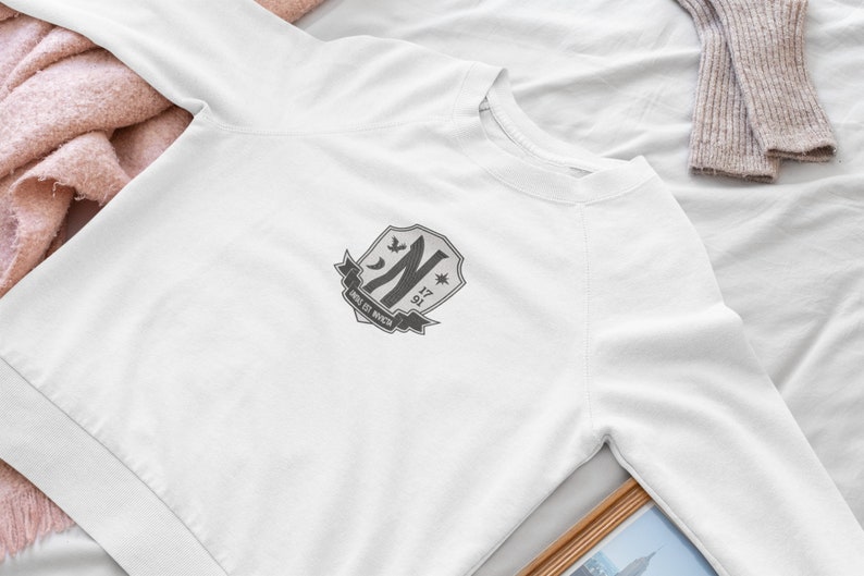 May include: White sweatshirt with a gray embroidered crest featuring a stylized letter "N", a star, and the text "NINE 17 91".