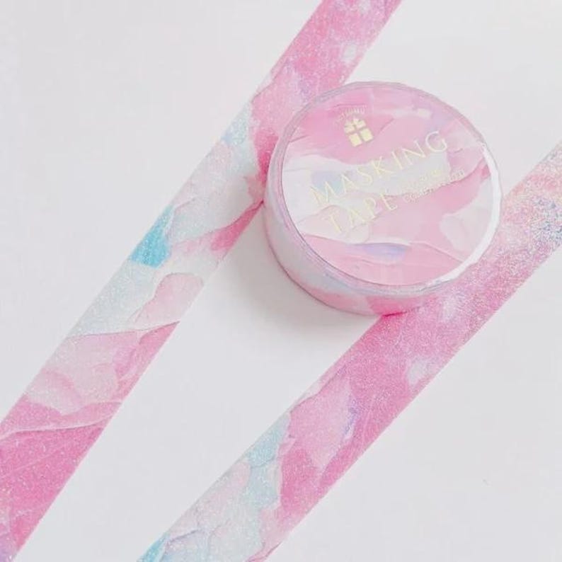 May include: A roll of masking tape and two strips of the same tape featuring a marbled pattern in shades of pink, white, and light blue with a subtle glittery finish. The circular roll label displays the text MASKING TAPE in gold lettering.
