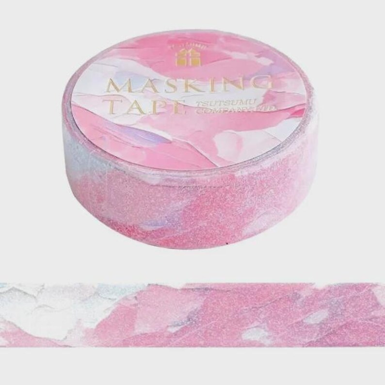 May include: A roll of masking tape with a pink, white, and light purple abstract watercolor pattern. The label on the top of the roll features the text MASKING TAPE and TSUTSUMU COMPANION in gold lettering, along with a small gold gift box icon.