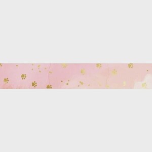 May include: A horizontal strip of decorative washi tape featuring a soft, light pink watercolor background. The surface is adorned with a repeating pattern of small, metallic gold paw prints scattered across the length of the tape.