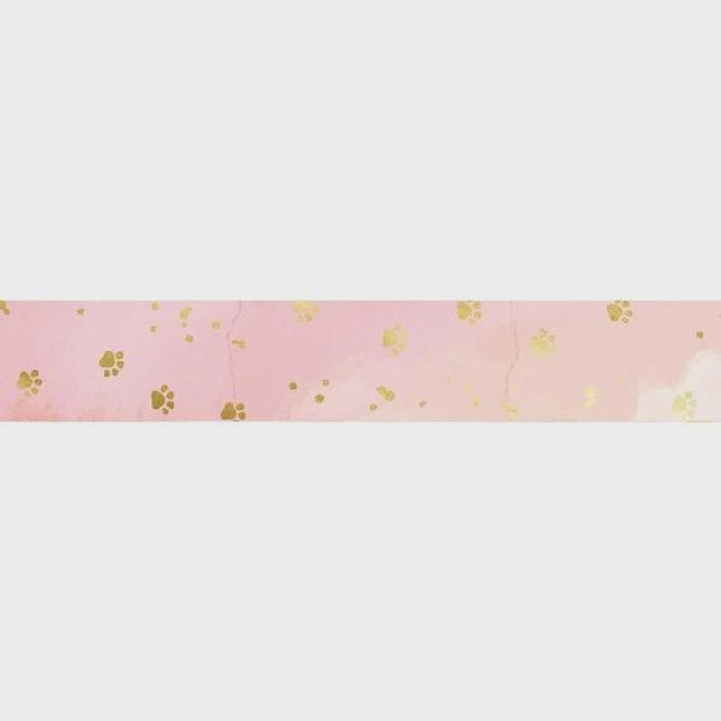 May include: A horizontal strip of decorative washi tape featuring a soft, light pink watercolor background. The surface is adorned with a repeating pattern of small, metallic gold paw prints scattered across the length of the tape.